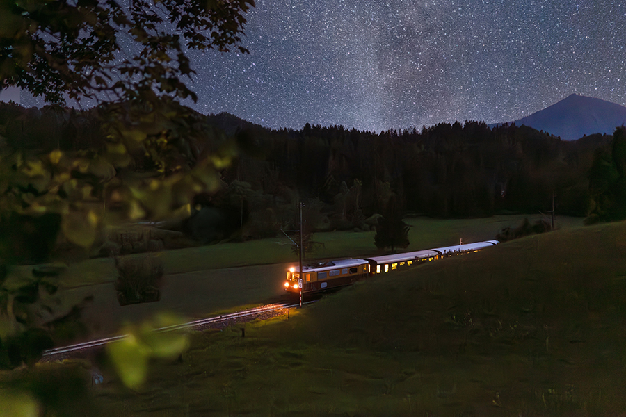 Ein Zug fährt nachts durch eine dunkle Landschaft unter einem sternenklaren Himmel mit vielen Sternen und einem Berg im Hintergrund