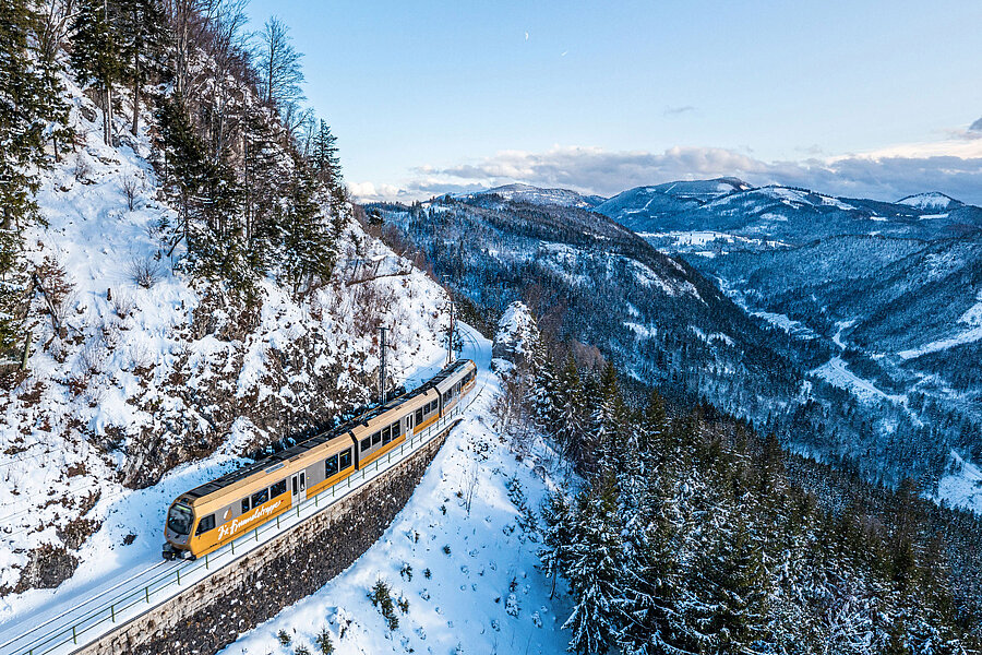 Mariazellerbahn im Winter Gelber Zug fährt auf verschneiter Bergstrecke entlang eines steilen Hangs mit Tannenbäumen und Blick auf bewaldete Berge