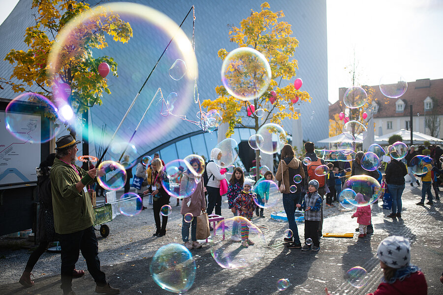 Ein Straßenkünstler erzeugt große Seifenblasen, während Kinder und Erwachsene an einem sonnigen Tag im Freien zuschauen und spielen, mit bunten Luftballons und Bäumen im Hintergrund.