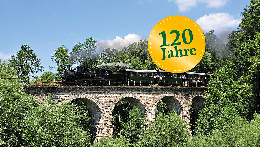 Rauchender Dampfzug fährt über ein Viadukt.