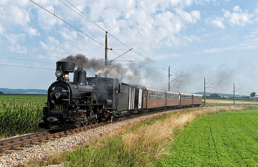 Der Dampfzug mit 4 Waggons fährt in einem Bogen. Aus der Lok kommt dunkler Rauch und die Landschaft im Hintergrund ist grün.