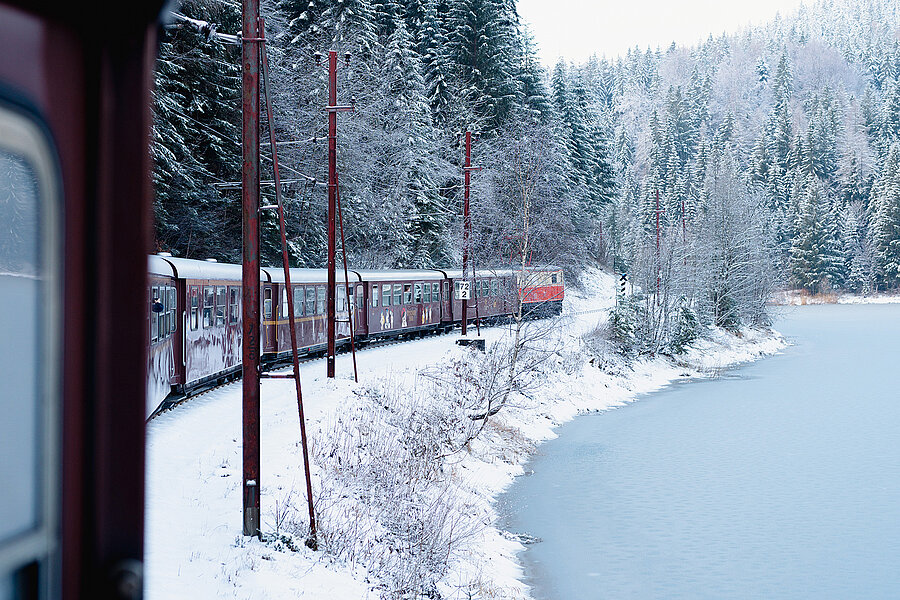 Ein roter Zug fährt entlang eines zugefrorenen Sees durch eine verschneite Winterlandschaft. Dichte, schneebedeckte Nadelwälder und Oberleitungen säumen die Bahnstrecke.