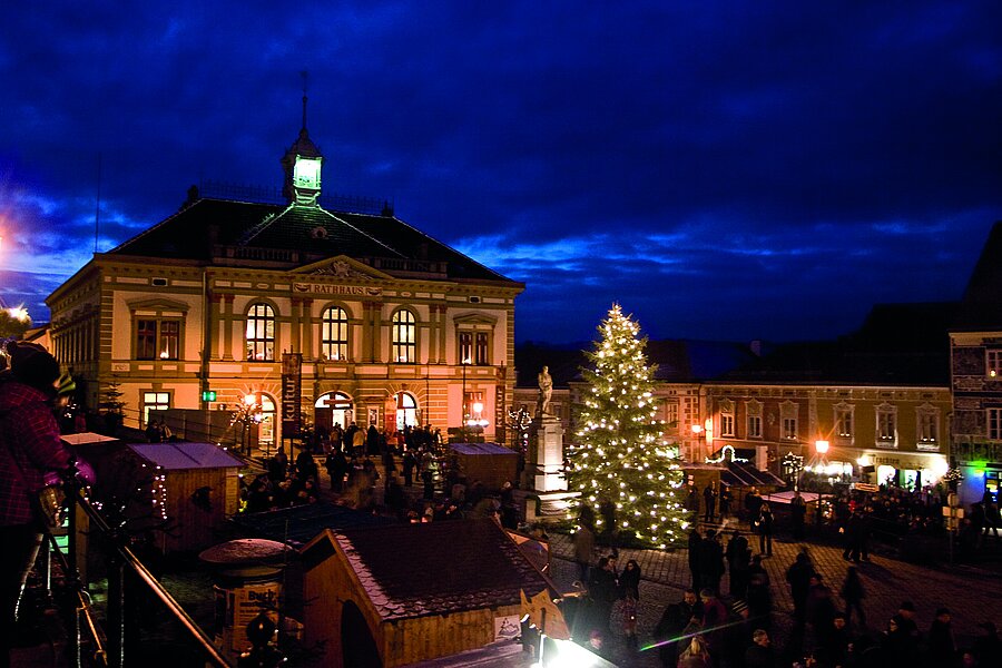 Weihnachtsmarkt in Weitra bei Nacht mit beleuchtetem Rathaus und großem Weihnachtsbaum, Menschen besuchen Stände