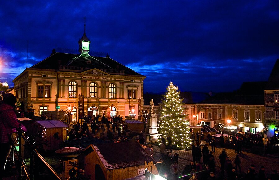 Weihnachtsmarkt in Weitra bei Nacht mit beleuchtetem Rathaus und großem Weihnachtsbaum, Menschen besuchen Stände