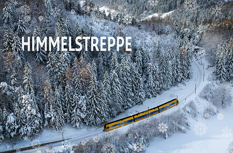 Die goldene Himmelstreppe fährt durch einen tief verschneiten Wald entlang der Mariazellerbahn. Weiße Schneeflocken und der Schriftzug „Himmelstreppe“ schmücken das winterliche Motiv.