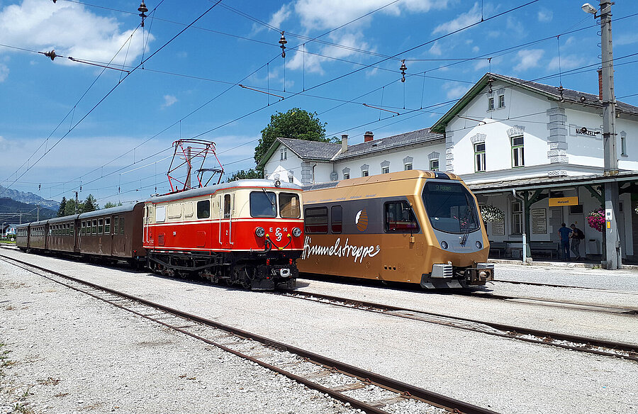 Historischer roter Zug und moderne goldene Himmelstreppe stehen nebeneinander am Bahnhof Mariazll.