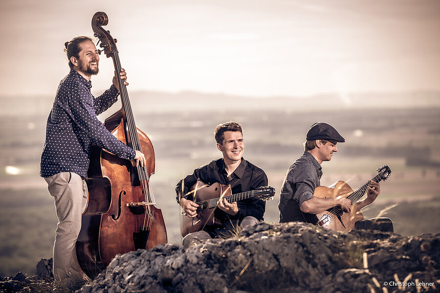 Drei Musiker spielen akustische Instrumente in einer natürlichen Felslandschaft mit weitem Ausblick über das Tal. Links steht ein Mann mit Dutt und Hemd am Kontrabass und lacht. In der Mitte sitzt ein Mann mit dunklem Hemd und spielt Gitarre, lächelnd in die Ferne blickend. Rechts sitzt ein weiterer Mann mit Schiebermütze und spielt ebenfalls Gitarre, konzentriert nach unten schauend. Die Szene ist warm beleuchtet mit weichem Hintergrund. 