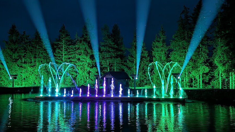 Nächtliche Wassershow mit türkis, grün und violett beleuchteten Fontänen auf einem See. Lichtstrahlen ragen in den Himmel, während Bäume und Wasser farbenfroh erstrahlen.