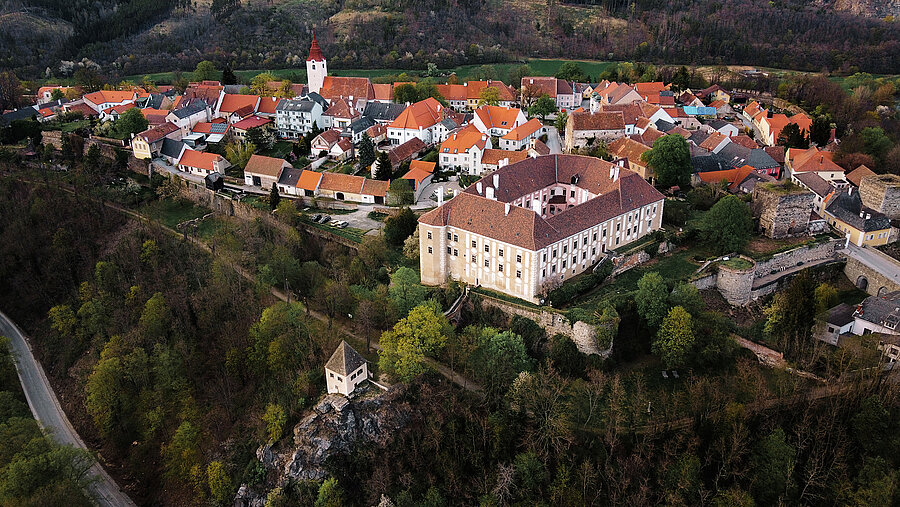 Luftaufnahme eines historischen Ortskerns mit rot gedeckten Häusern, umgeben von grüner Hügellandschaft. Im Vordergrund ein großes Schloss mit Innenhof auf einem Hügel, darunter ein kleiner Aussichtspavillon auf einem Felsen. Rechts vom Schloss erstreckt sich die Altstadt mit engen Gassen und einer Kirche mit rotem Turm. Mauern und Reste alter Befestigungen umgeben das Areal.