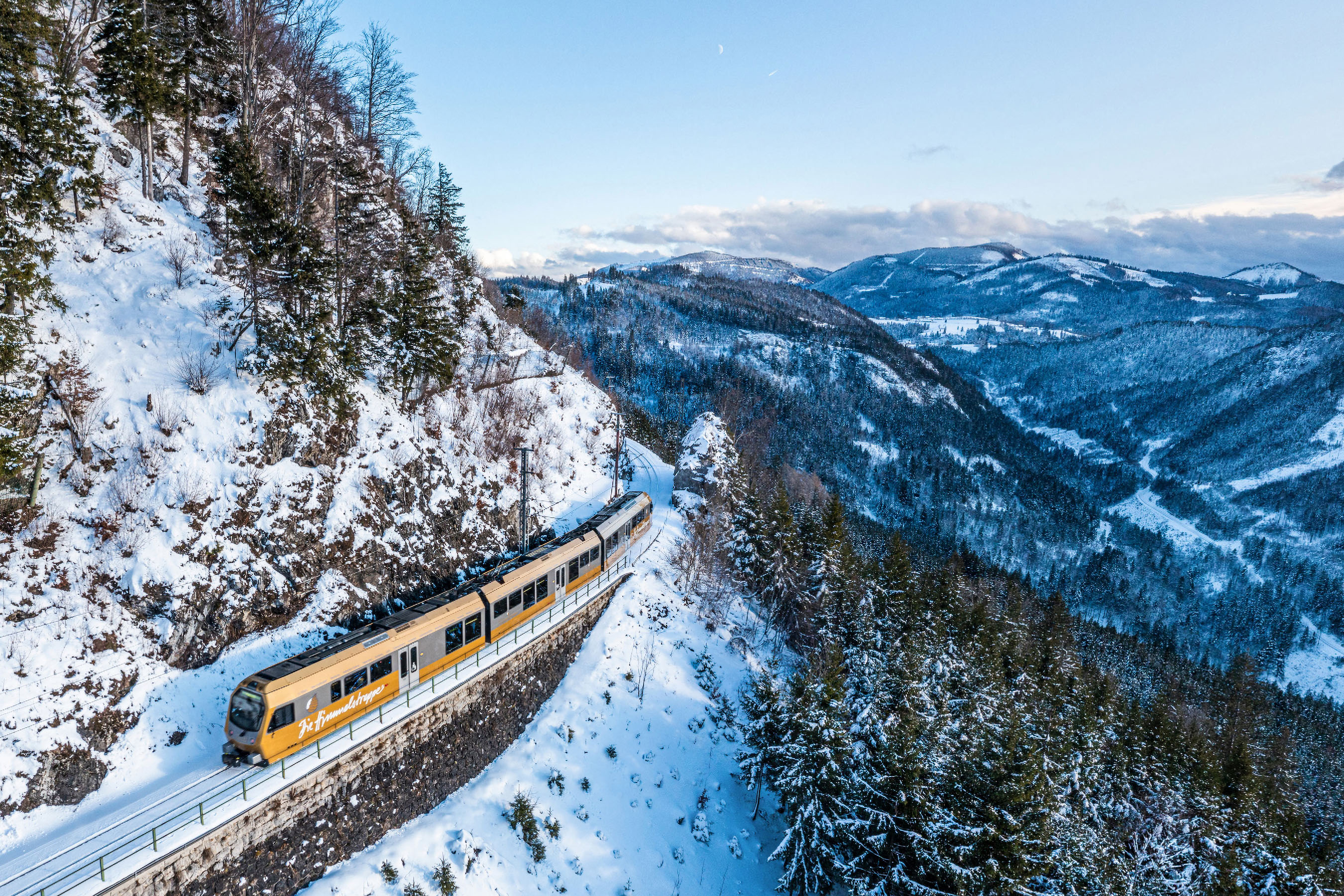 Gelber Zug fährt auf verschneiter Bergstrecke entlang eines steilen Hangs mit Tannenbäumen und Blick auf bewaldete Berge